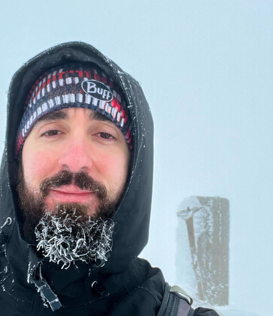 Close-up selfie of a bearded man in a hood and beanie, his beard covered in ice crystals, with snowy fog and a frost-covered trail sign behind him.